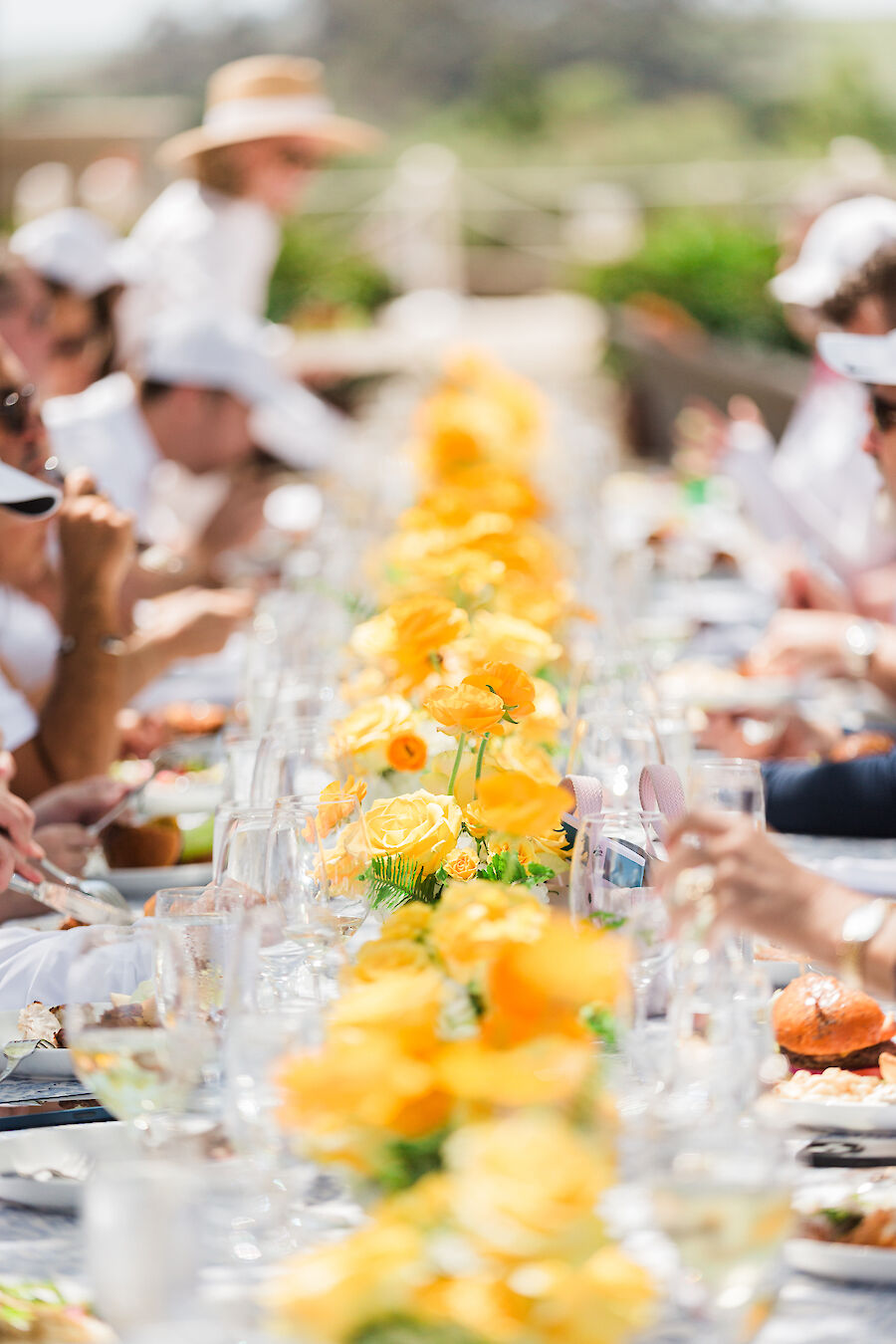 A long table with a centerpiece of yellow flowers, surrounded by people dining outdoors, creates a festive and elegant atmosphere.