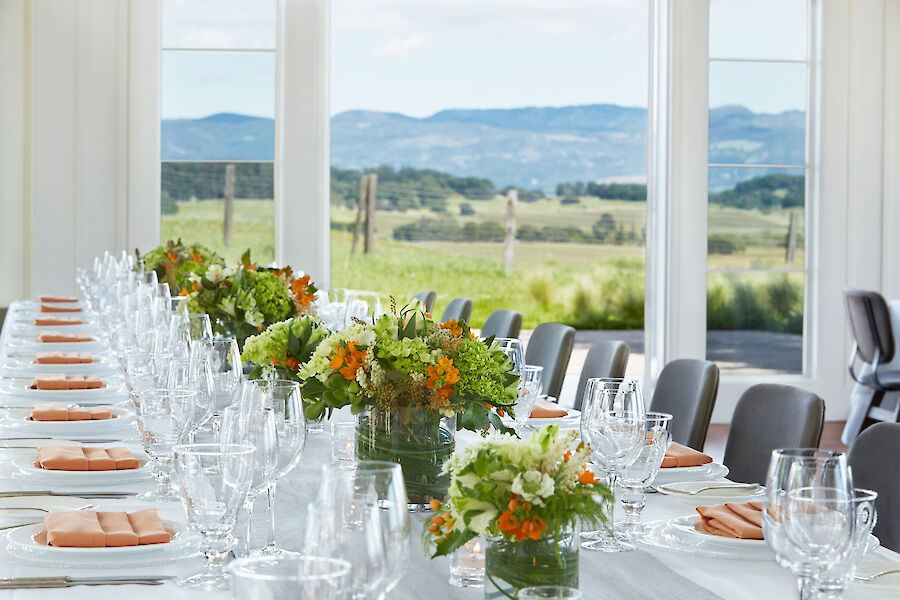 A long dining table is elegantly set with glassware, plates, and floral centerpieces, overlooking a scenic landscape through large windows.