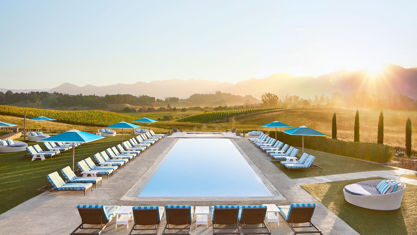A serene outdoor pool surrounded by lounge chairs and umbrellas, set against a backdrop of vineyards and mountains at sunrise or sunset.