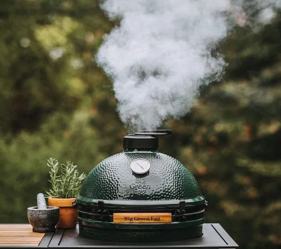 A green ceramic grill or smoker emits smoke outdoors; plants and mortar with pestle nearby on a wooden surface.