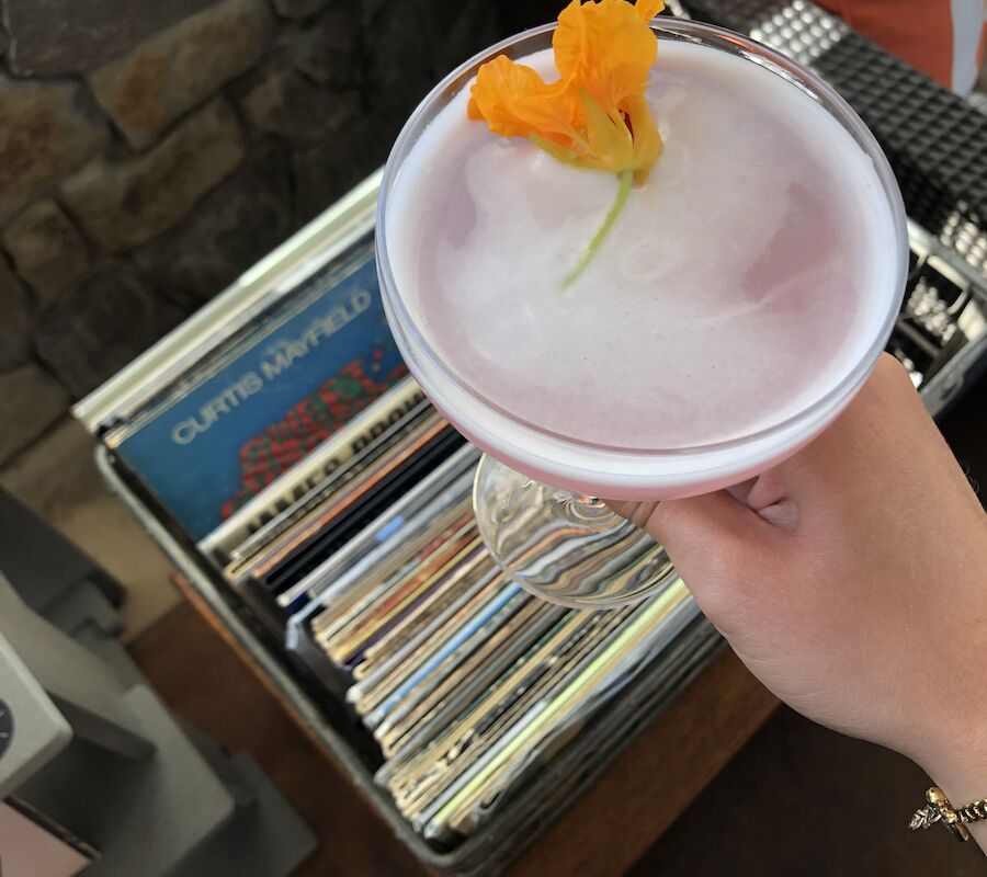 A person holds a cocktail with an orange flower garnish, next to a stack of vinyl records.