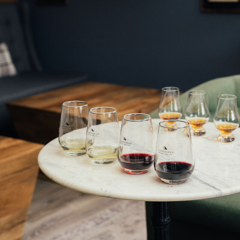 A round table with glasses of wine and whiskey in a cozy seating area, featuring green and gray chairs.