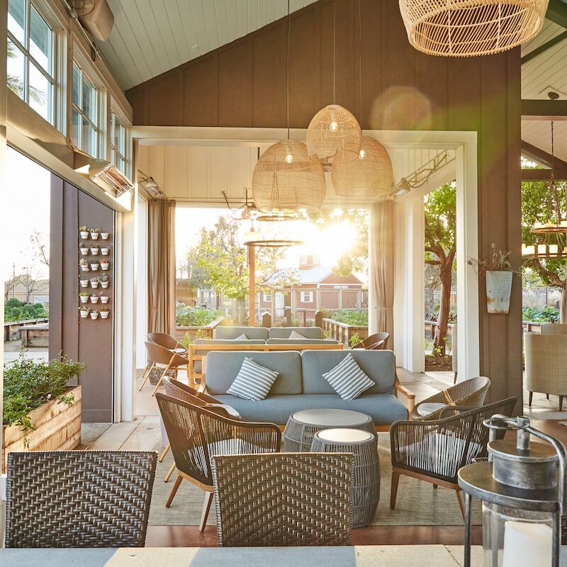 A sunlit patio with wicker furniture, cozy seating, hanging lamps, and large windows opening to a garden view.