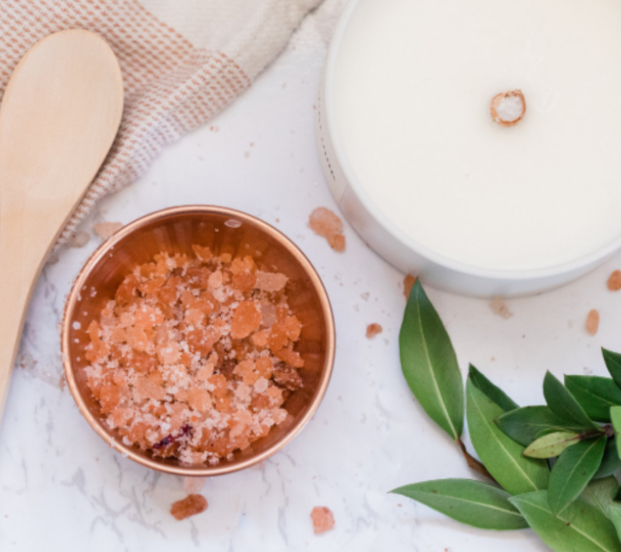 The image shows a wooden spoon, a bowl of pink bath salts, a white candle, and some green leaves on a marble surface.