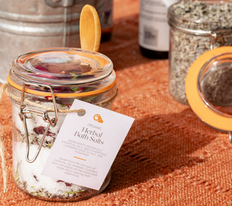 A jar of herbal bath salts with a wooden spoon on top, labeled, sits on an orange surface near other jars.
