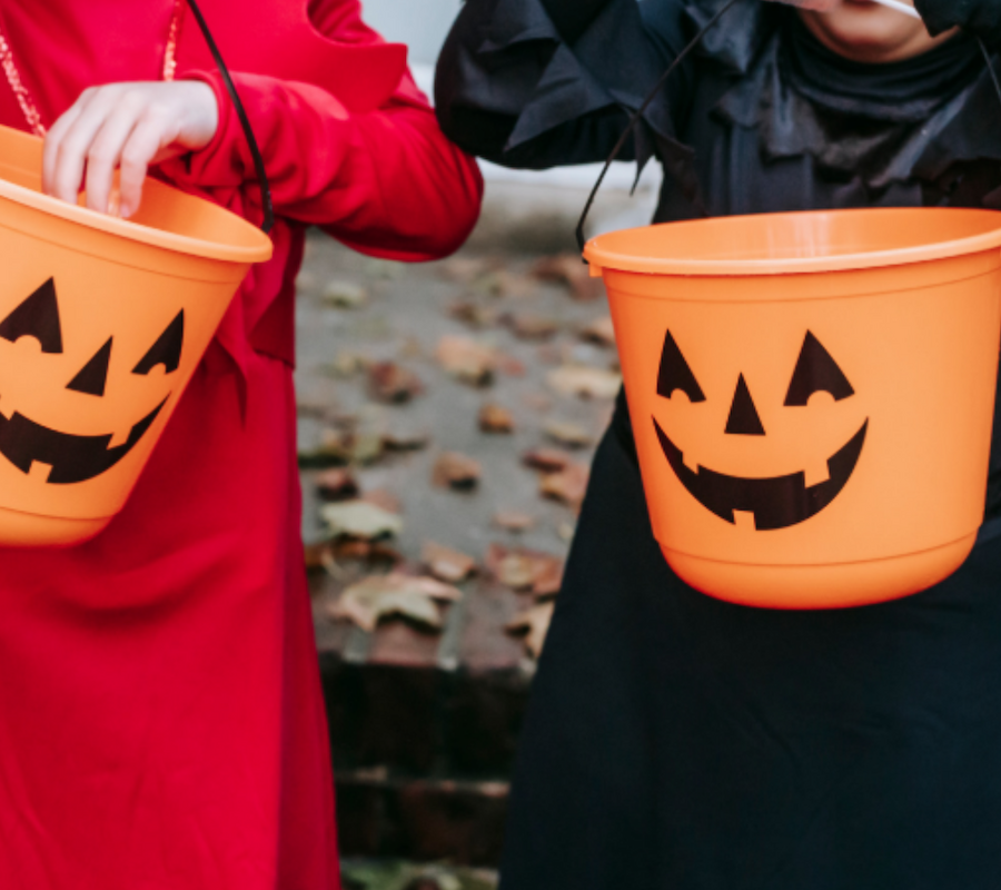 Two people in costumes hold orange buckets with jack-o'-lantern faces, likely for Halloween trick-or-treating.