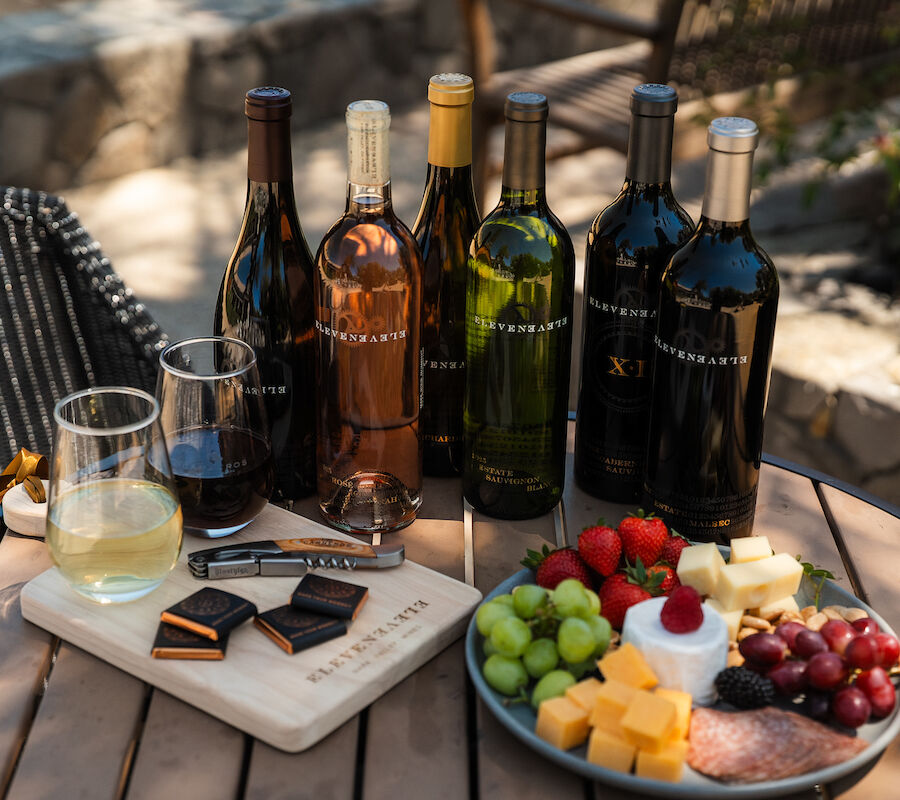 A variety of wine bottles, glasses, cheese, fruit, and snacks are elegantly arranged on an outdoor table.