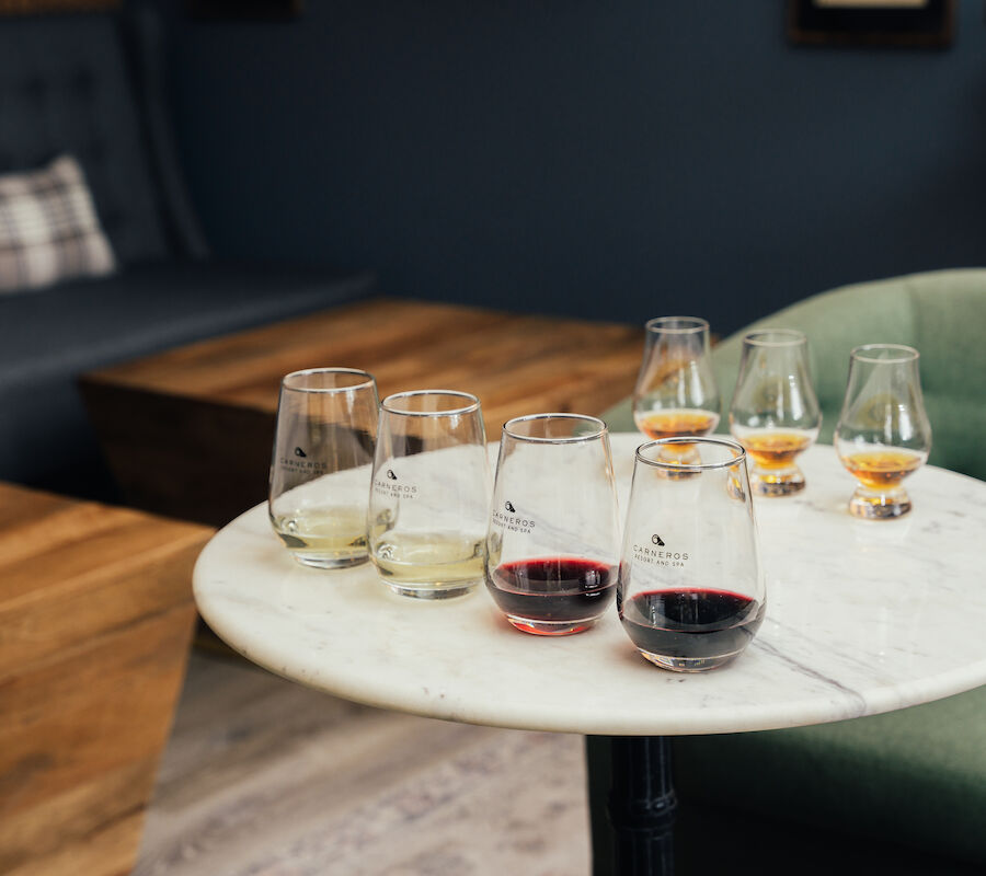 A round table with six glasses of wine and whiskey, set in a cozy room with a couch and green chair nearby.
