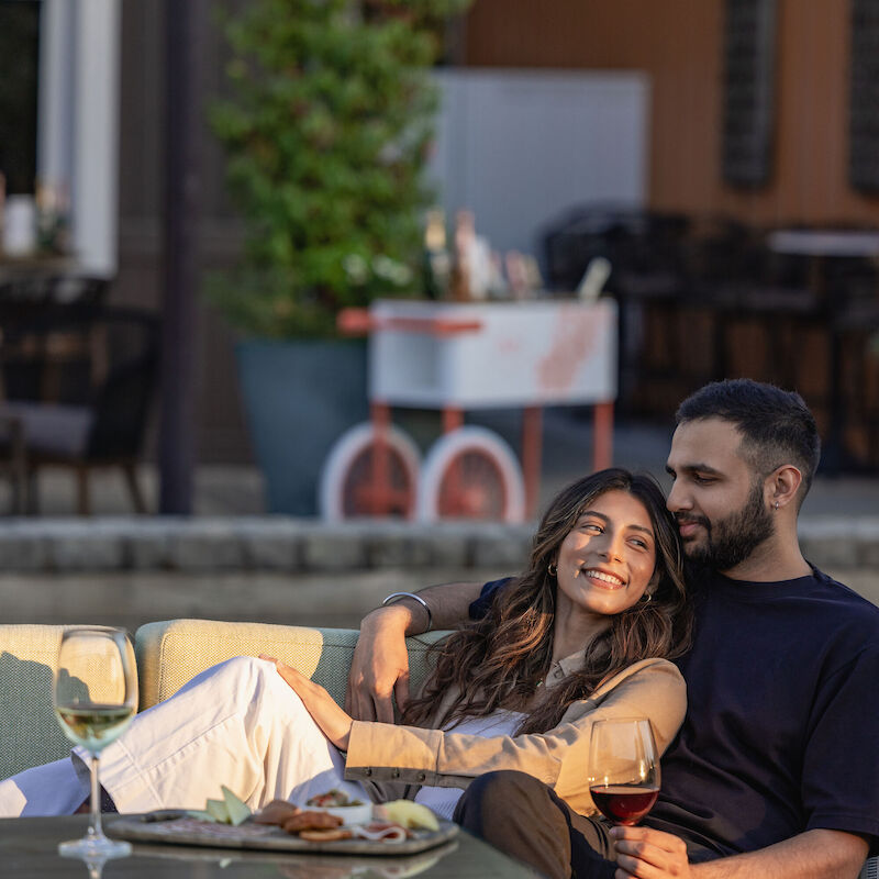 A couple is relaxing on a couch outdoors, enjoying drinks and appetizers, with a cozy vibe and a blurred background.