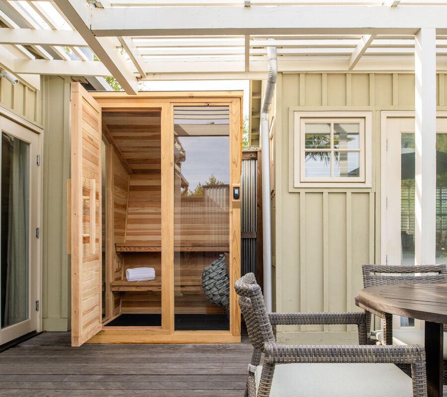 This image shows an outdoor patio with a wooden sauna, wicker chairs, and a wooden table under a pergola.