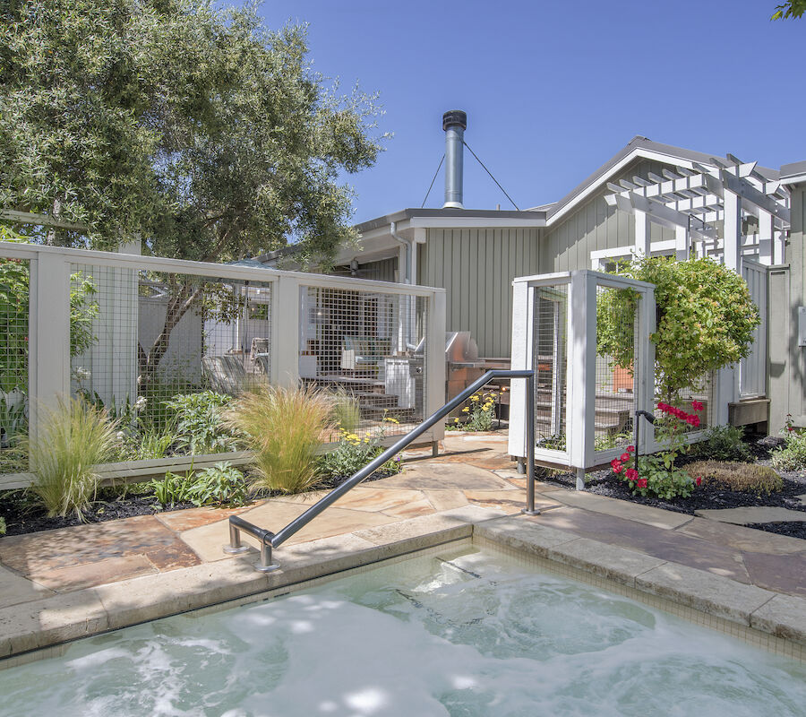 A serene outdoor area with a hot tub, surrounded by greenery, a cozy building, and a clear blue sky in the background.