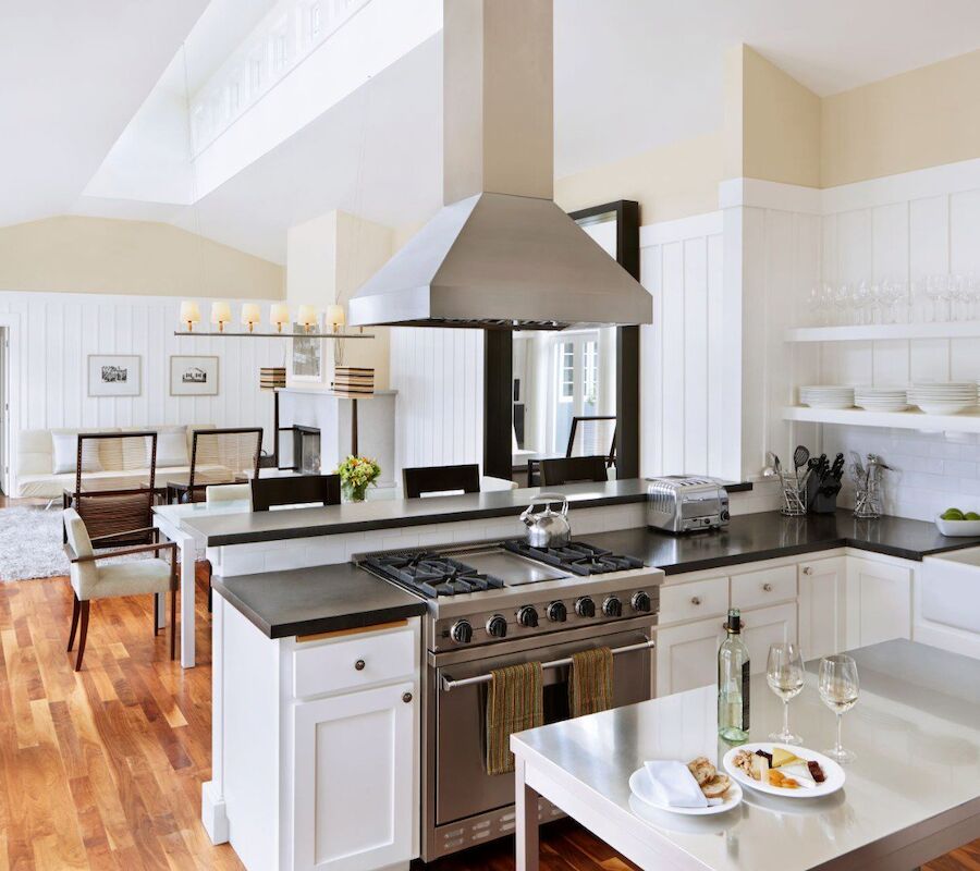 A modern kitchen with white cabinetry, stainless steel appliances, and a dining table set. The space is bright and airy with wooden flooring.