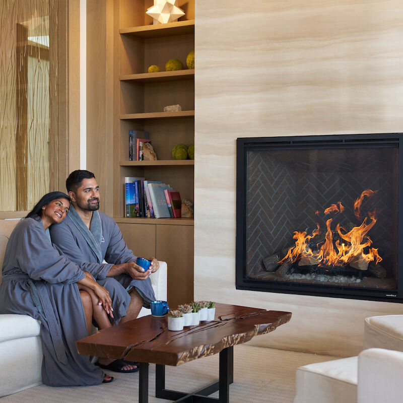 A couple in robes sits by a modern fireplace, surrounded by shelves and a wooden table, creating a cozy atmosphere.