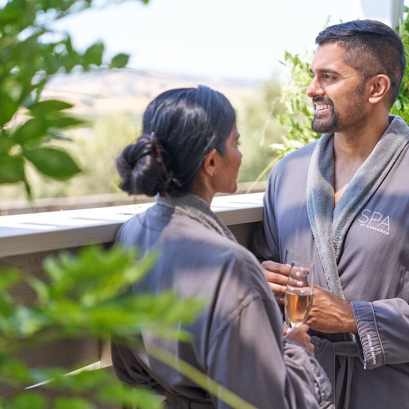 Two people in spa robes are enjoying a conversation outdoors, with one holding a drink.