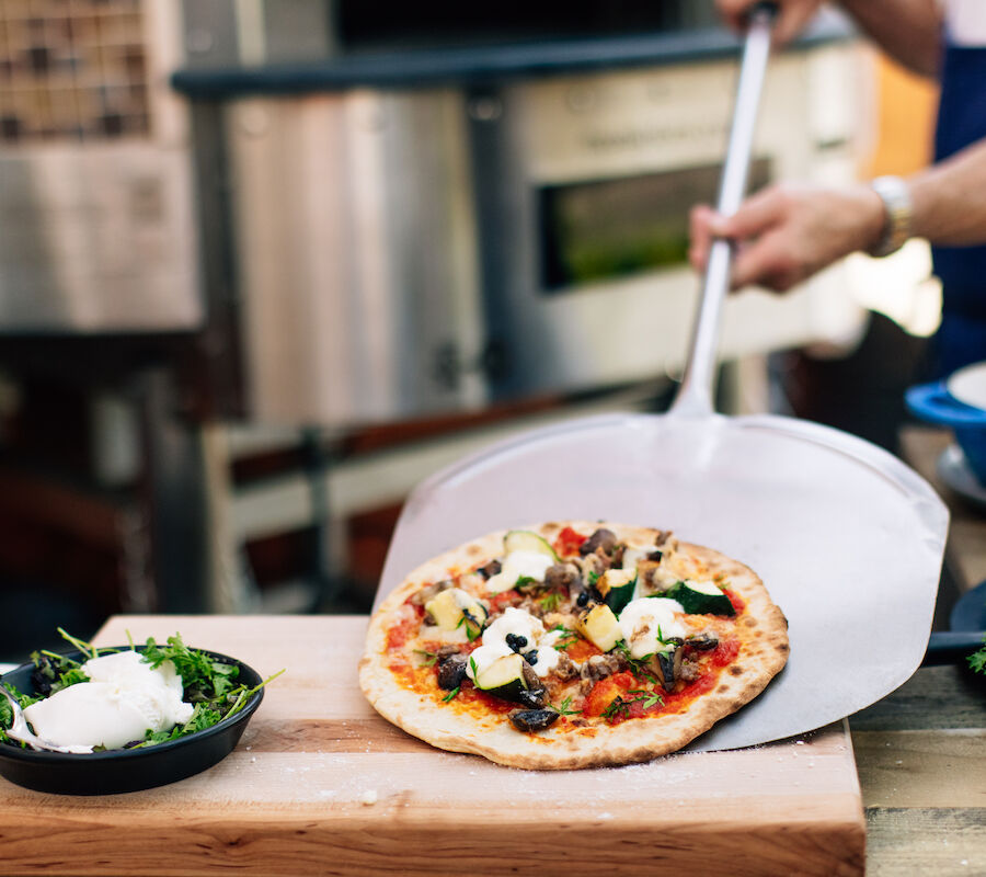 A person is using a pizza peel to place a freshly baked pizza on a wooden surface, surrounded by ingredients and greenery.