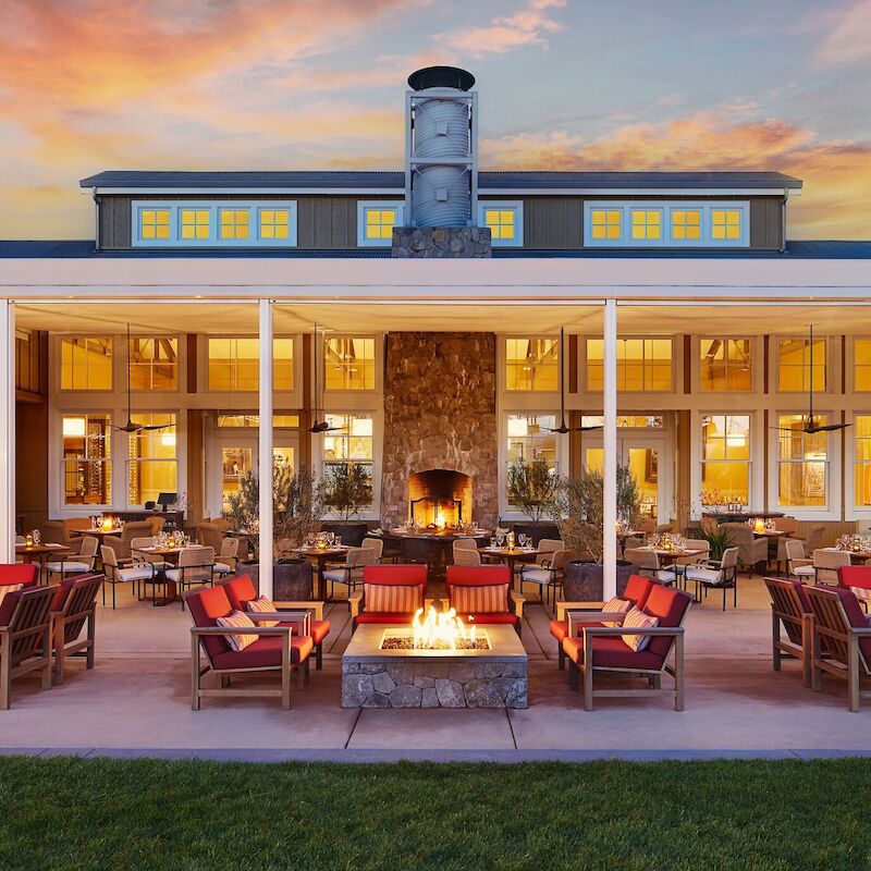 Outdoor seating area with fire pits and chairs, in front of a building with large windows and a chimney at sunset.