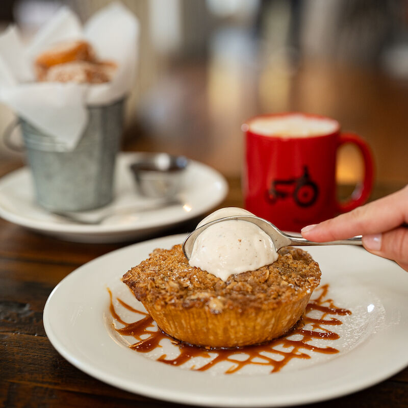A dessert topped with ice cream sits on a white plate, next to coffee and pastries in the background, on a wooden table.