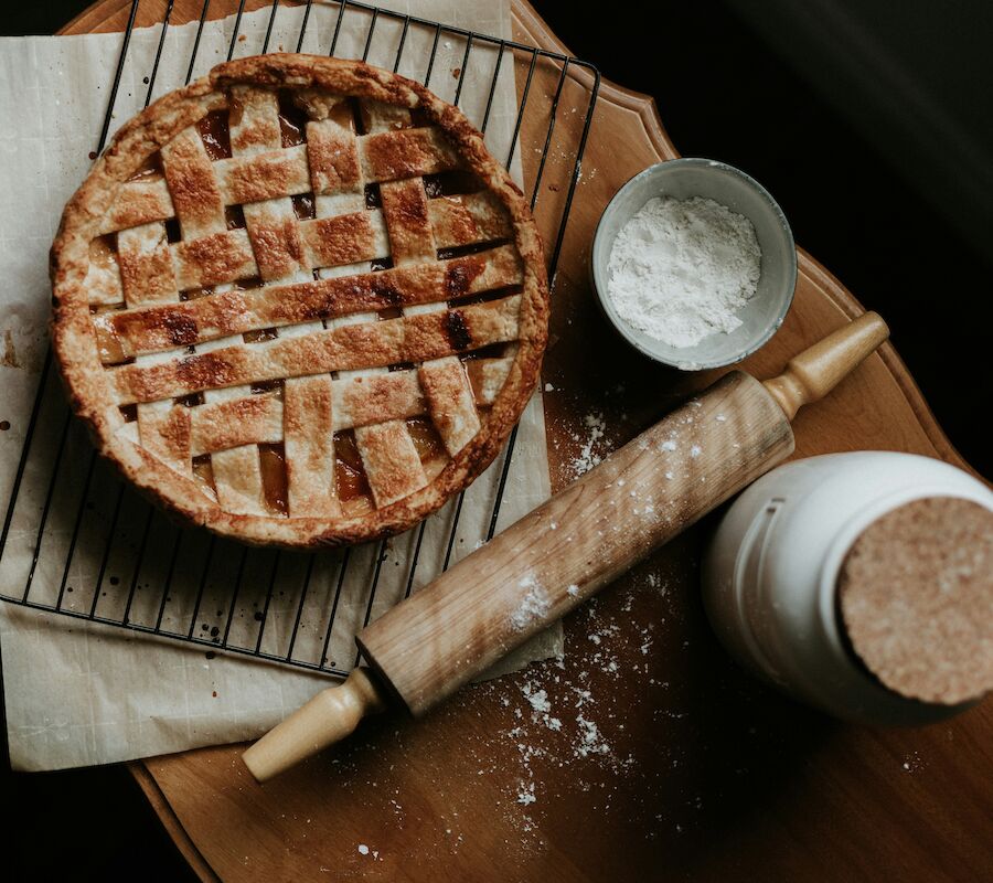 A pie with a lattice crust on a cooling rack, a bowl of flour, a rolling pin, and a jar on a wooden surface.