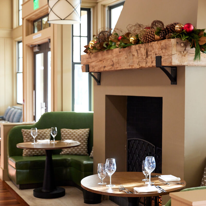 A cozy restaurant interior with green booths, round tables set with wine glasses, and a decorated fireplace mantel.