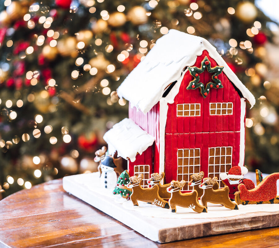 A festive gingerbread house with reindeer and a sleigh is displayed on a table, with a Christmas tree in the background.