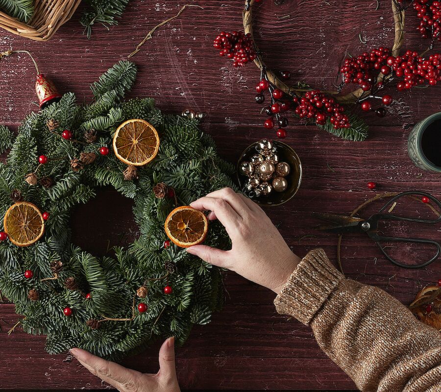 A person decorates a wreath with dried oranges and berries on a wooden table. Nearby are crafting tools, greenery, and a cup.