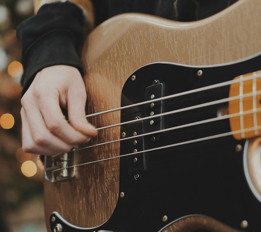 A person is playing a bass guitar with a light wood finish. There are blurred lights in the background, possibly from a Christmas tree.