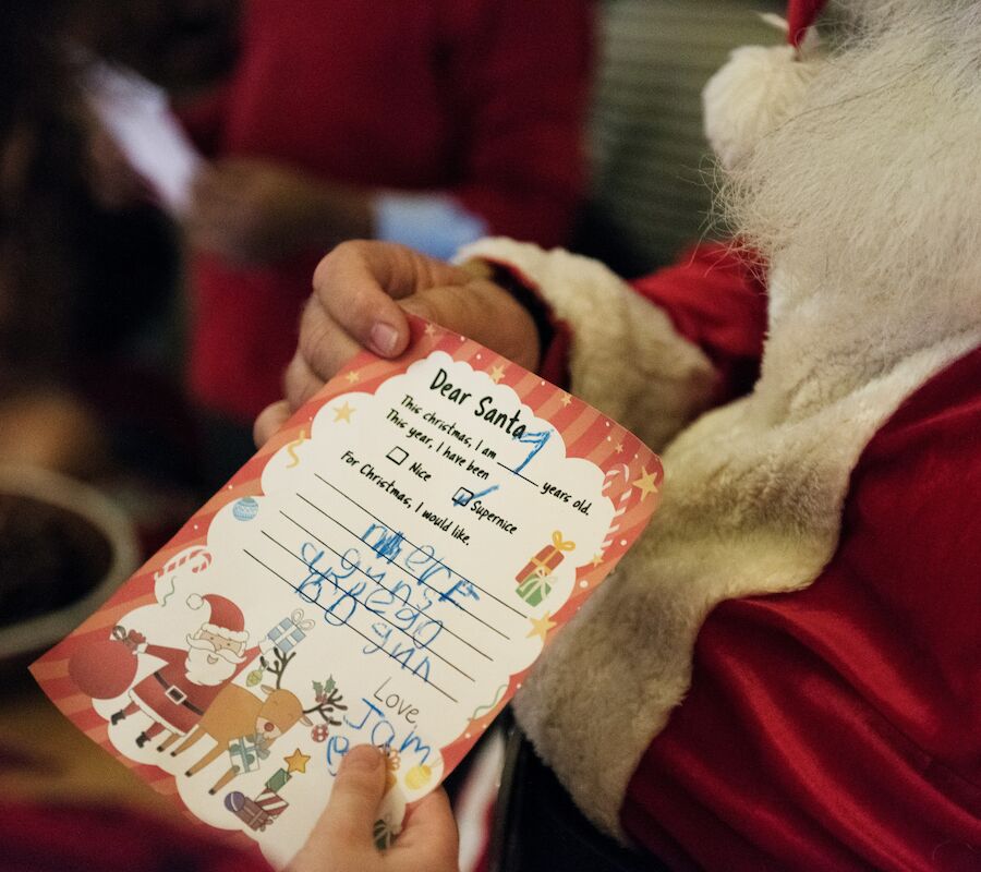 A person in a Santa outfit holds a festive letter addressed to Santa, written in childlike handwriting, with Christmas-themed graphics.